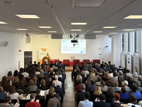 Im Auditorium des Grimm-Zentrum sind circa 150 Personen von hinten zu sehen, die auf Stühlen sitzen und der Referentin Gabriele Zöllner zuhören, die die Konferenz eröffnet. Gabriele Zöllner steht dabei auf einr Bühne vor einem Podest, im Hintergrund sind ihre Vortragsfolien an die Wand projiziert.