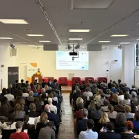 Im Auditorium des Grimm-Zentrum sind circa 150 Personen von hinten zu sehen, die auf Stühlen sitzen und der Referentin Gabriele Zöllner zuhören, die die Konferenz eröffnet. Gabriele Zöllner steht dabei auf einr Bühne vor einem Podest, im Hintergrund sind ihre Vortragsfolien an die Wand projiziert.